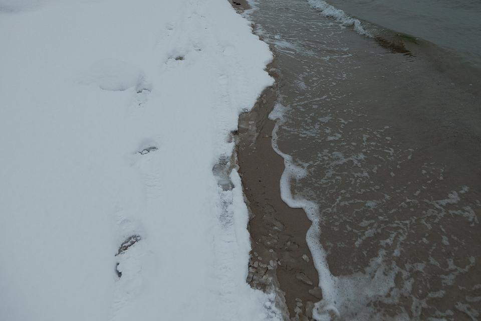 The beach is split in two, snow on the side water on the other. Footprints cut through the white, deliberate but not hurried.The waves reach them, hesitate, then pull back.