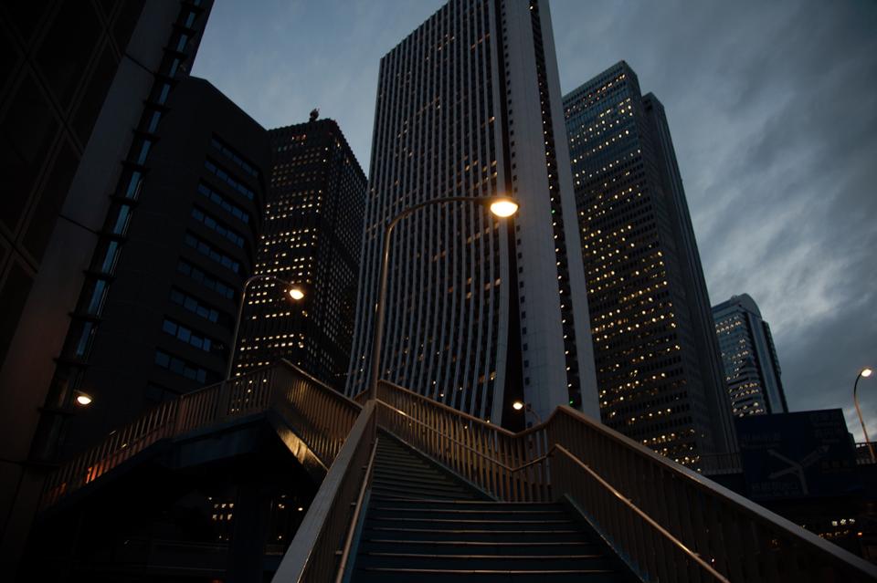 The staircase cuts diagonally through the frame, directing the eye upward.
Towers rise beyond it, their interiors lit in quiet repetition.
Streetlights punctuate the path.
The city feels structured, efficient, and distant.