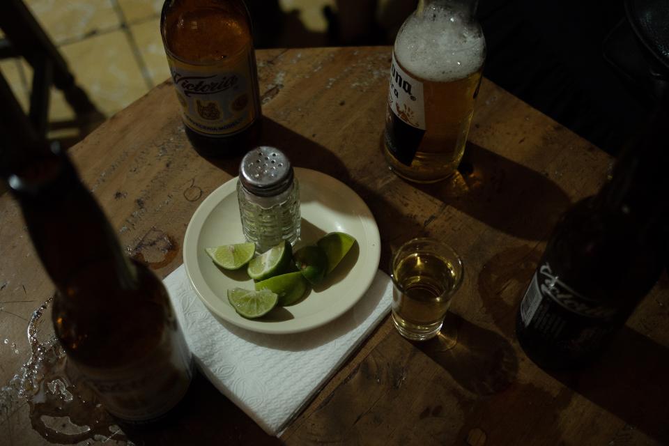 Rain presses against the street outside.
Green neon hums over the room.
Daylight dissolves into electric shadow.
Beer and tequila move in quiet ritual.
Salt.
Lime.
A burn that answers the rain.