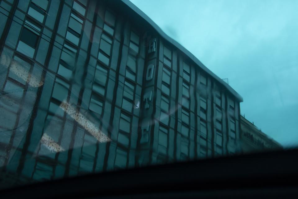 Rain bends the building into wavering lines.
Letters blur but remain.
The glass does not block the city.
It distorts it.