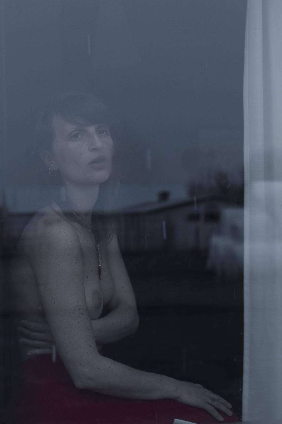 Light filters through a thin layer of reflection on the window.
A woman sits just inside, bare from the waist up, her posture closed.
The exterior landscape overlays her figure.
The image holds both sides at once.