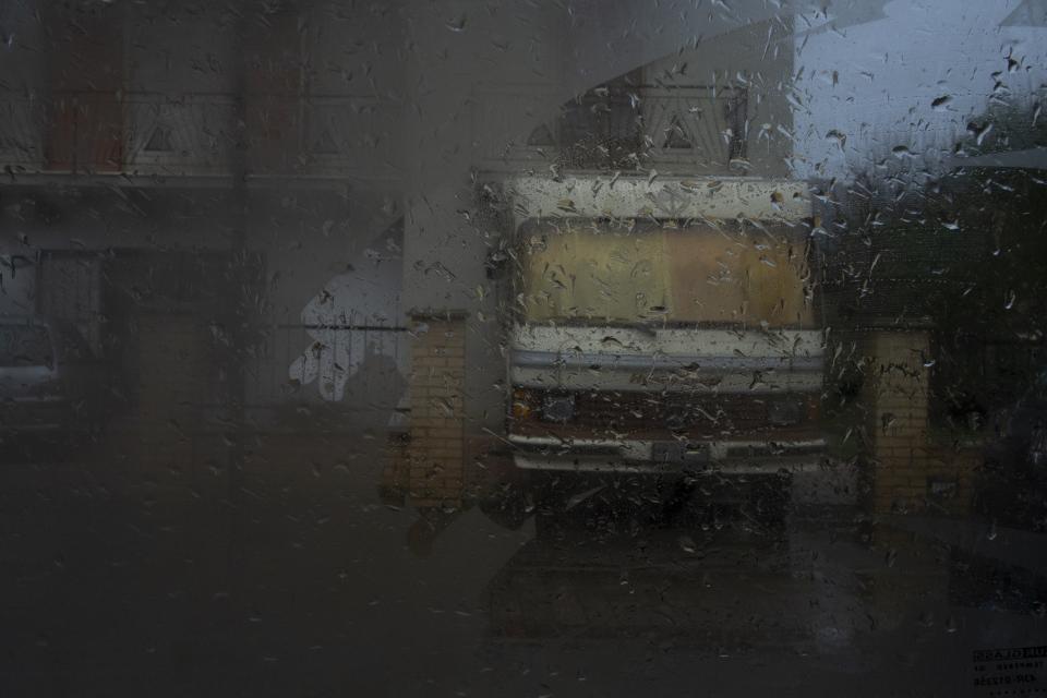 Rain covers the glass in irregular patterns.
Droplets gather, hesitate, then slide downward.
Outside, a truck is parked directly in front of the building.
Its headlights are off.
Its shape remains intact but softened by water.
The window becomes a second surface.
The world beyond is visible, yet distorted.
The window functions as an emotional boundary.
The rain acts as interference.
The world outside becomes something that cannot be touched.