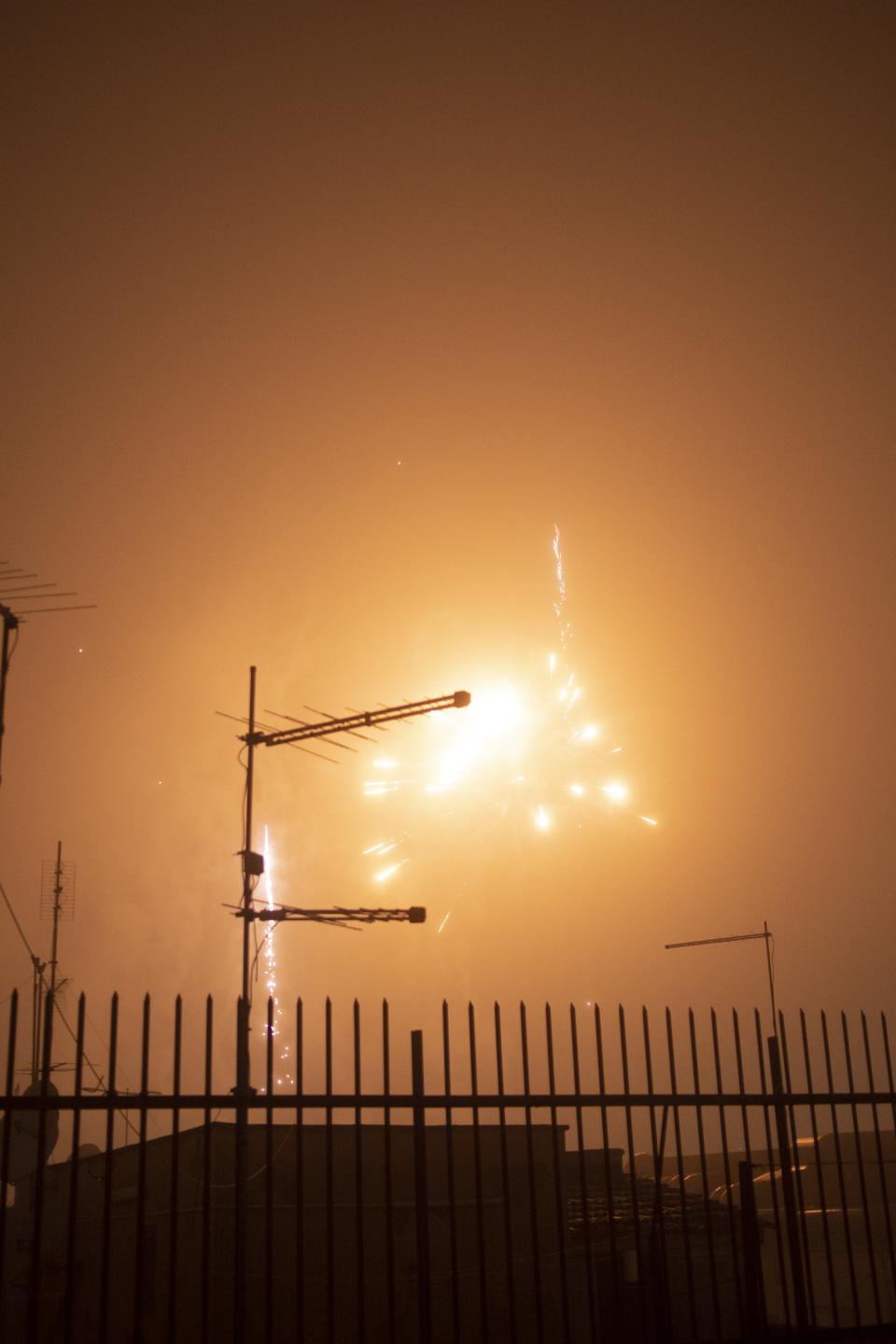 Fireworks bloom in an orange sky.
Antennas slice the light into rigid lines.
Sparks flare and vanish.
The fence stays still.