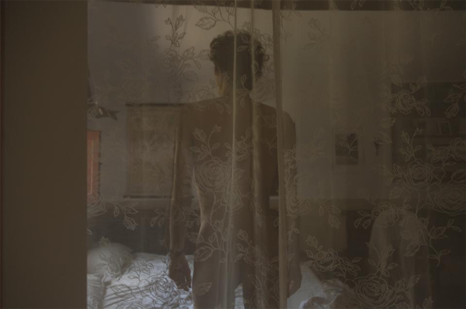 Soft amber light fills the room.
A figure stands still at the edge of the bed.
A sheer curtain embroidered with roses veils the body.
Details blur into silhouette.
