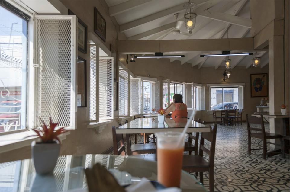 Bright light spills through open windows.
Street sounds blur at the edges.
A woman sits alone with the afternoon.
The camera remains still —
a silent observer,
half-hidden behind the edge of a glass.