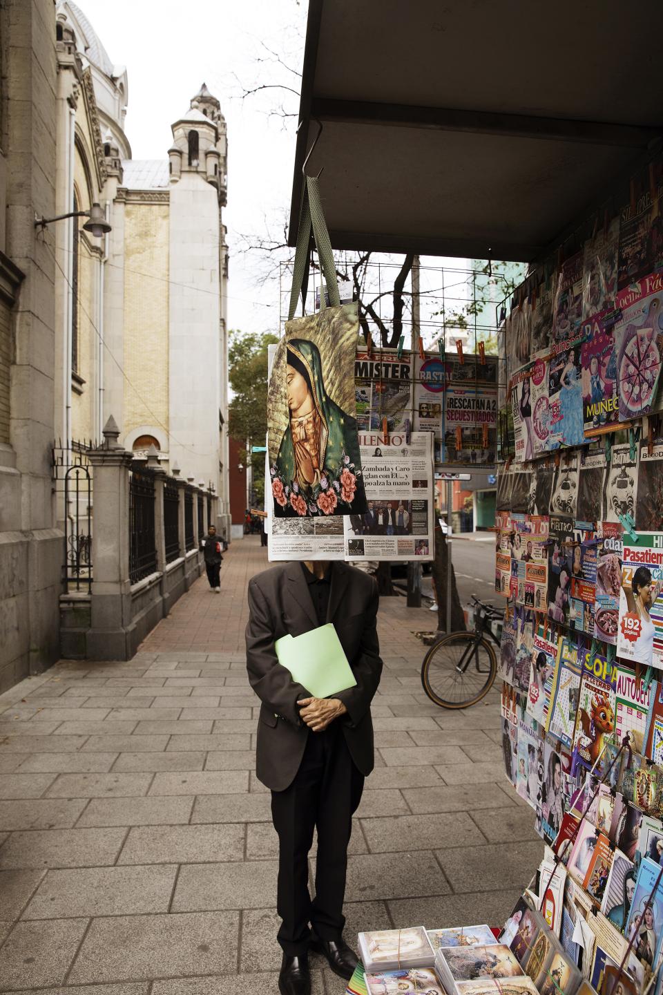 City sidewalk under pale light.
Magazines crowd the frame with color and noise.
A suited figure stands still.
The face replaced by an image of devotion.
Sacred and mundane overlap.
The street keeps moving.
